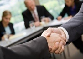 Two people shaking hands with three people at a desk writing in the background
